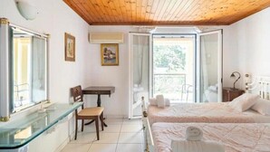 Une chambre avec un plafond en bois, un bureau en verre, un lit avec une literie blanche et une vue sur l’extérieur par une porte ouverte.