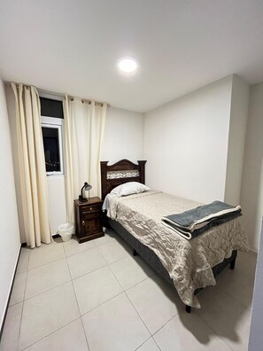 A bedroom with a bed, a nightstand, a window with curtains, a radiator, and a tiled floor.