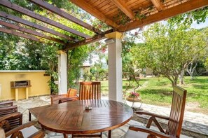 Una pérgola de madera con una mesa redonda y sillas, rodeada de follaje y árboles.