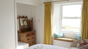 Ein Schlafzimmer mit einem Bett, einer Holzkommode, einem Spiegel, einem Fenster mit gelben Vorhängen und Blick ins Freie.