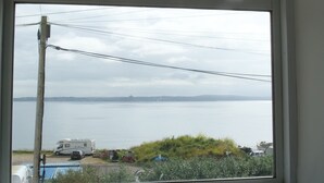 Ein Fenster mit Blick auf ruhiges Gewässer, einen abgestellten Wohnmobil und eine entfernte Küstenlinie.