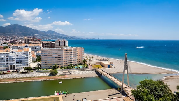 Un paisaje urbano costero con un puente atirantado destacado, una playa y un puerto deportivo.