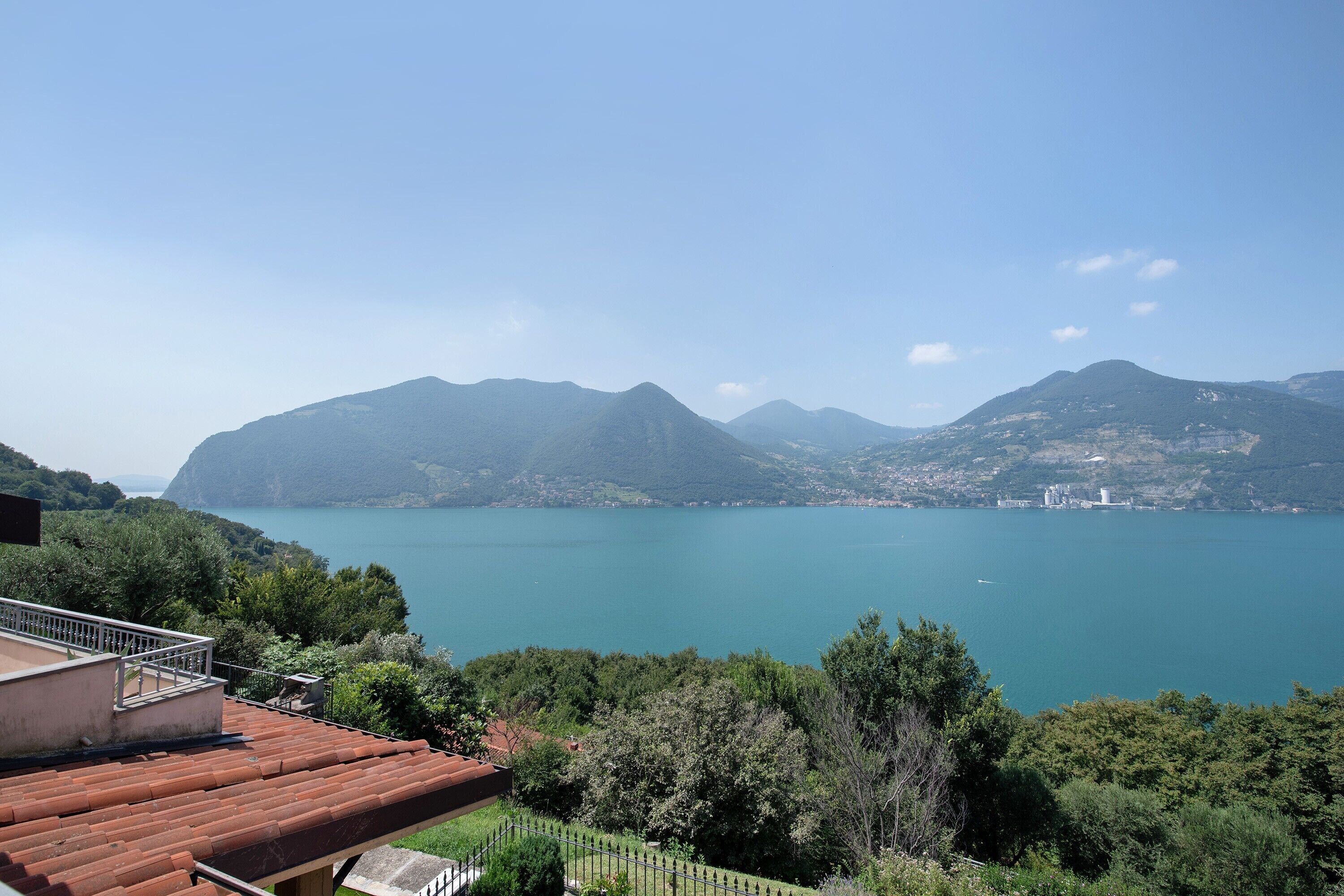 A view of a lake with mountains in the background, a building with a red roof, and a fence.