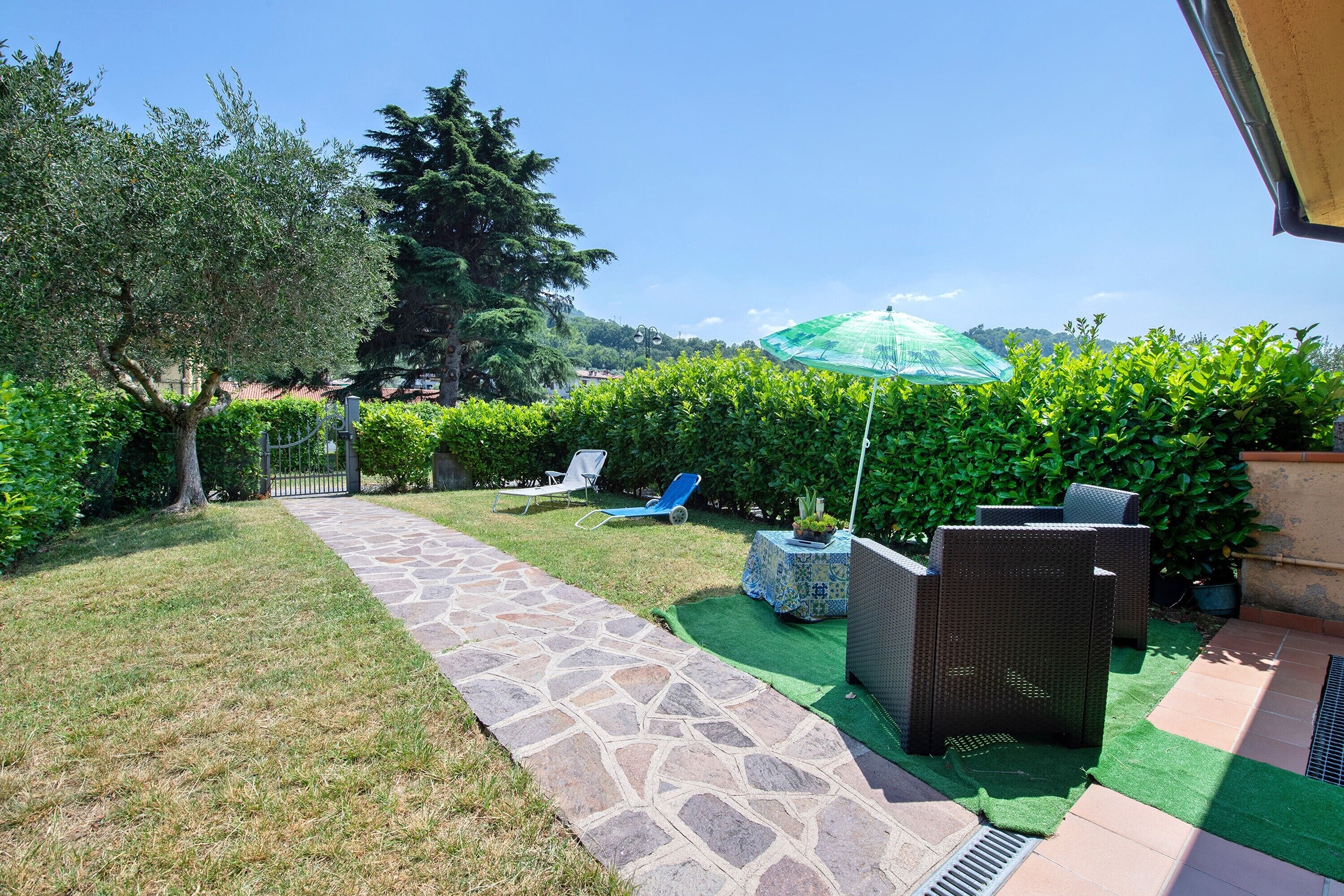 A stone pathway leading to outdoor seating with a green umbrella, surrounded by well-maintained hedges and trees.