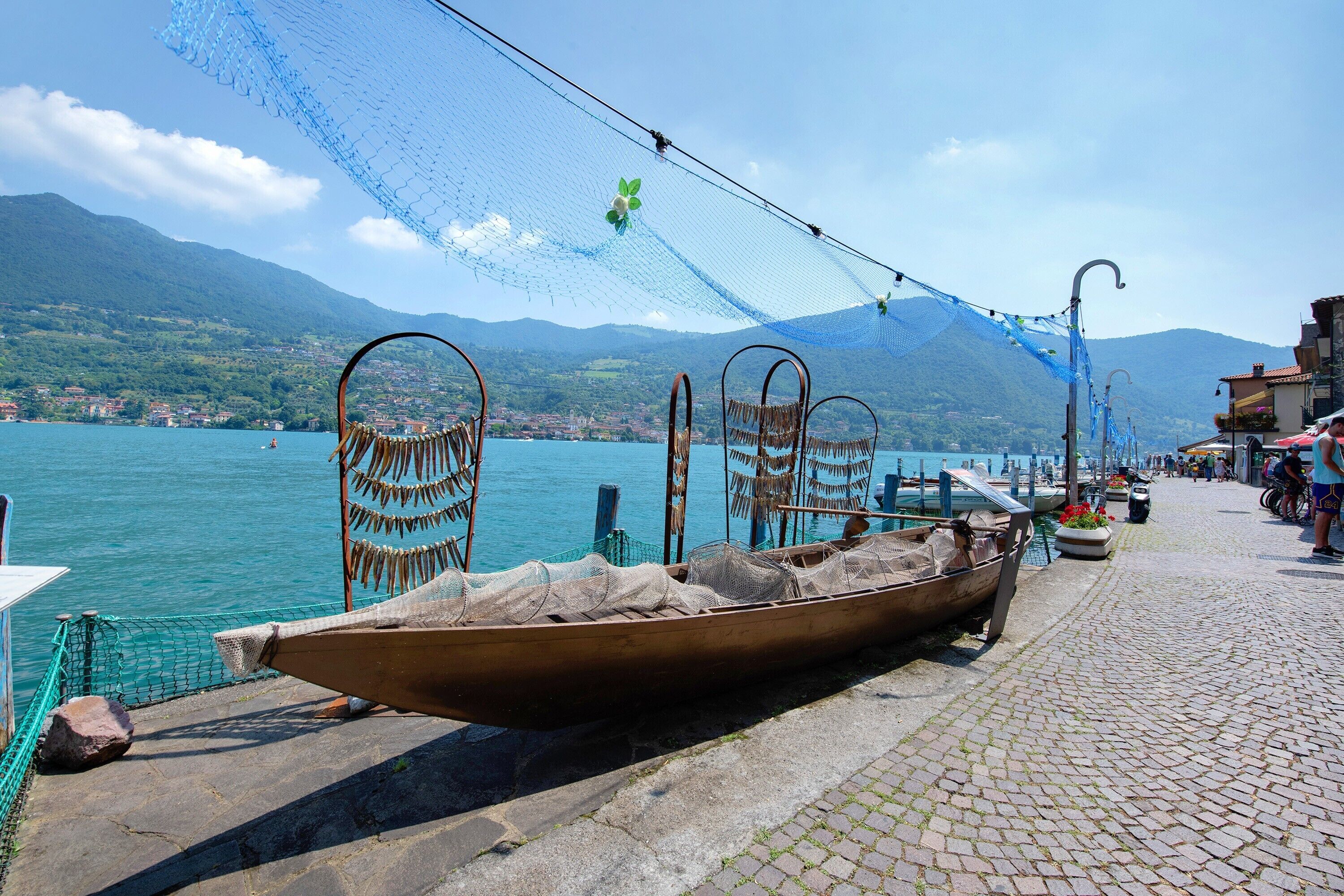A wooden boat docked by a cobblestone waterfront with a netted area overhead and mountains in the background.