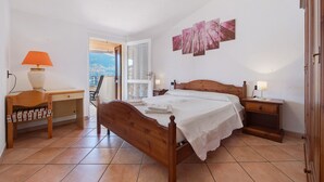 A bedroom with a wooden bed, a desk, a chair, a nightstand, and a view through an open door.