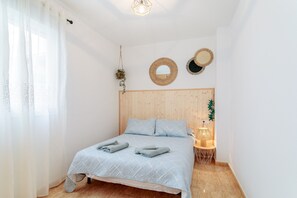 Une chambre avec une tête de lit en bois, un lit recouvert d’une couvre-lit à motifs, deux miroirs muraux, une plante suspendue et une lampe de chevet.