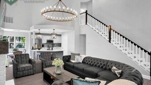 A spacious living area with a large sectional sofa, two armchairs, a coffee table, and a chandelier. A kitchen is visible in the background with white cabinets and a central island. - Luxury 4BR Beach House on the Sand (Redington Beach)