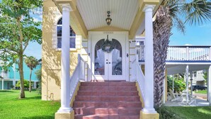 A hotel entrance with a covered porch, steps leading to a door, and a view of the surrounding area including a palm tree and another building. - Luxury 4BR Beach House on the Sand (Redington Beach)