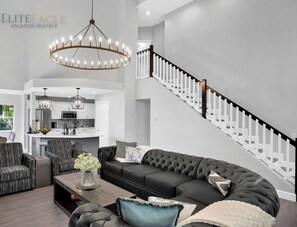 A spacious living area with a large sectional sofa, two armchairs, a coffee table, and a chandelier. A kitchen is visible in the background with white cabinets and a central island.
