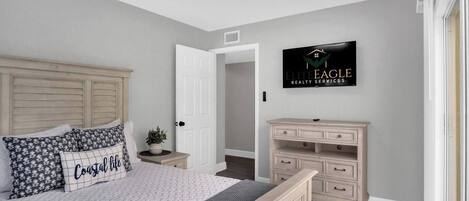 A bedroom with a bed, a nightstand, a dresser, a TV, and a door to another room.