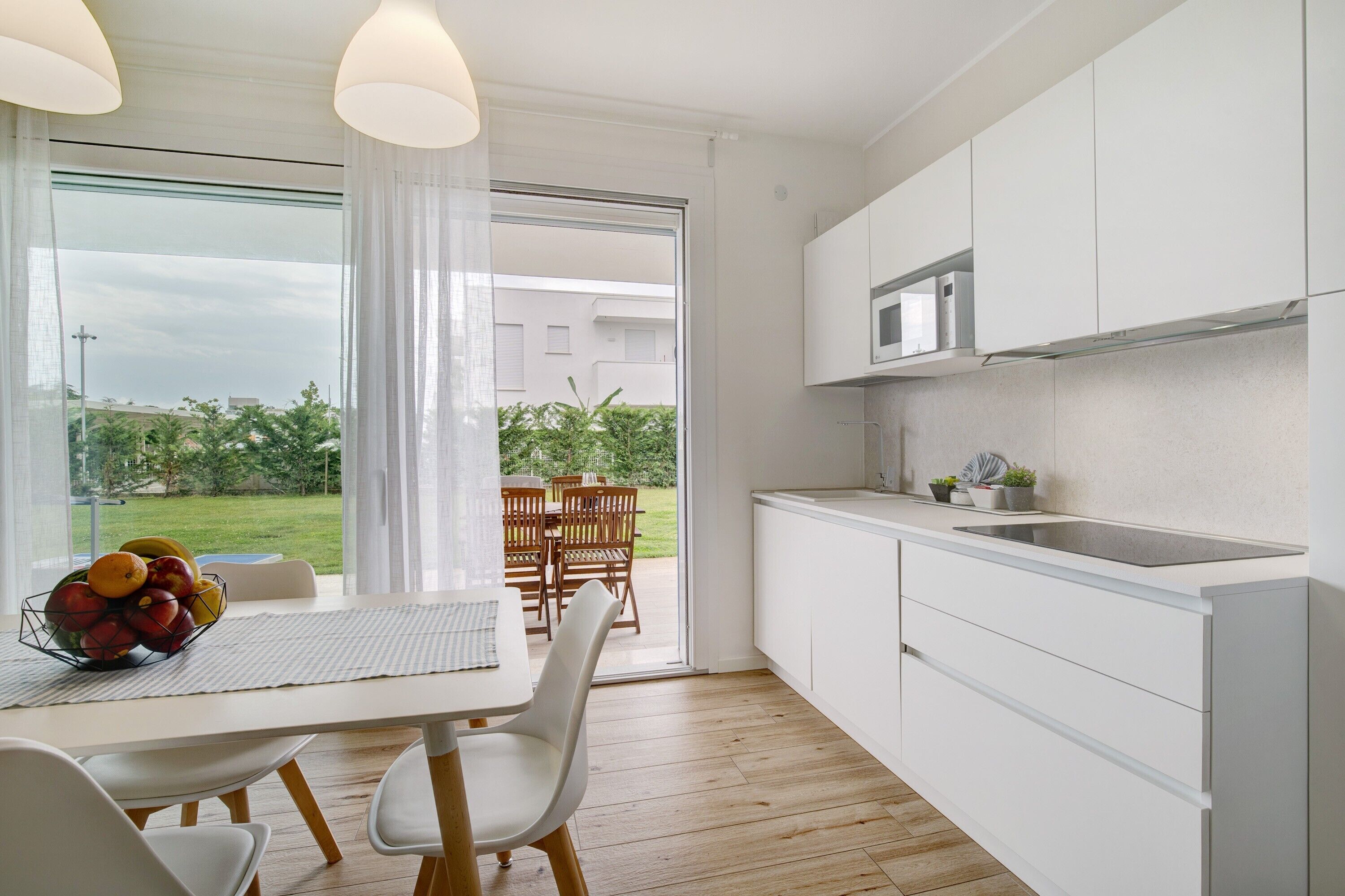 A modern kitchen with white cabinets, a microwave, and a sink. There is a dining table with chairs and a fruit bowl.