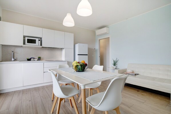 Une cuisine moderne avec des meubles blancs, un four à micro-ondes et un réfrigérateur. On y trouve une table à manger avec des chaises et un panier de fruits.