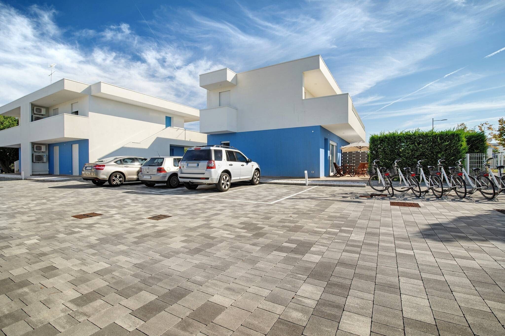 Un espace de stationnement où sont garées plusieurs voitures et des vélos, un bâtiment à la façade bleue et blanche, et un ciel dégagé.