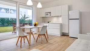 Eine moderne Küche mit weißen Schränken, einer Mikrowelle und einem Kühlschrank. Ein Essbereich mit Tisch und Stühlen lädt ein, und ein großes Fenster bietet Blick ins Freie.