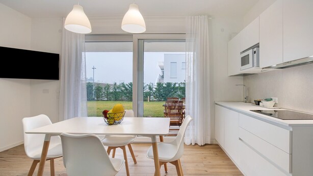 Eine moderne Küche mit weißen Schränken, ein Essbereich mit weißem Tisch und Stühlen sowie ein großes Fenster mit Blick auf Grünflächen.