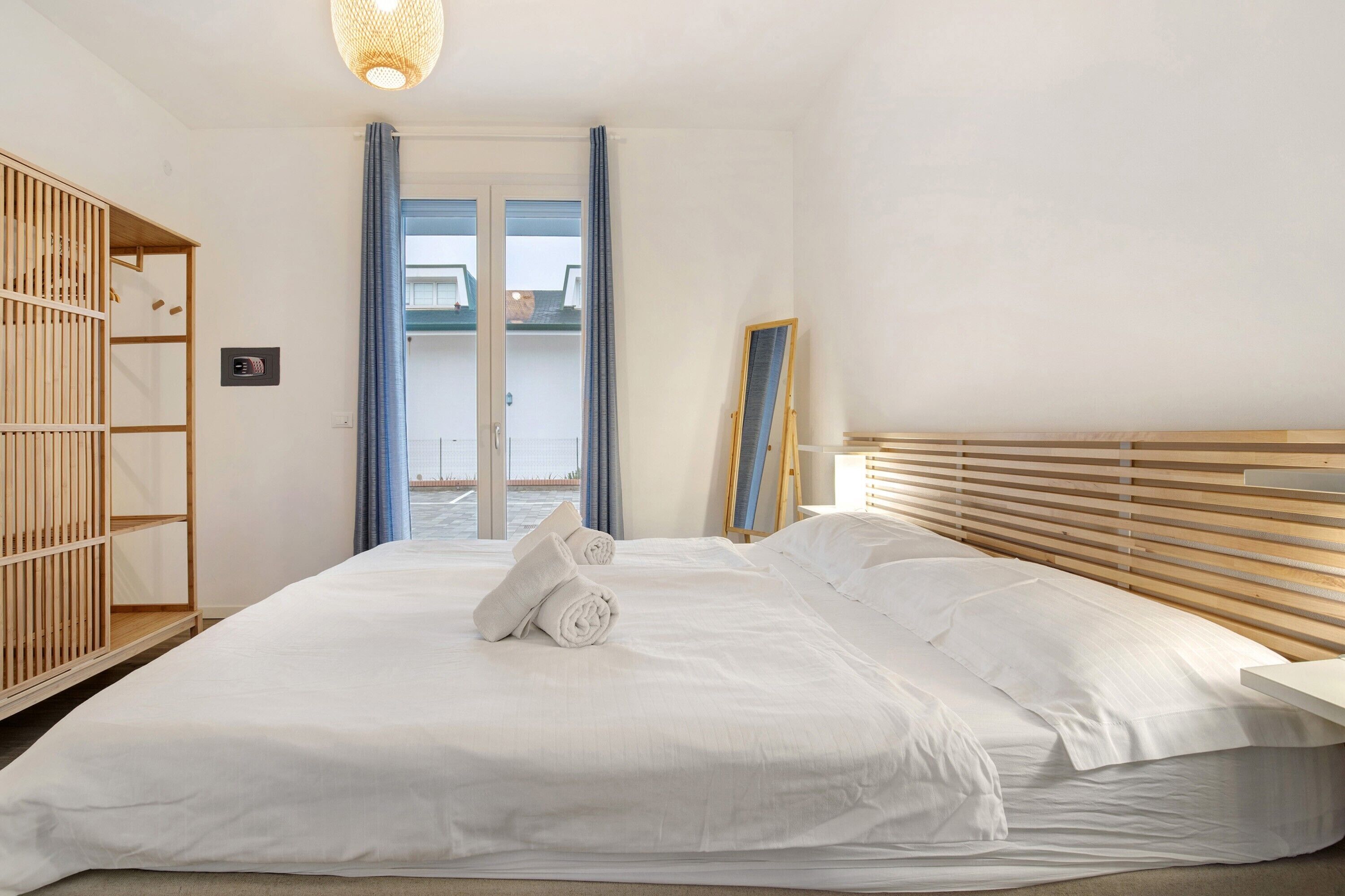 A hotel room with a bed, a wooden headboard, a wardrobe, a mirror, and a view of the sea through the door.