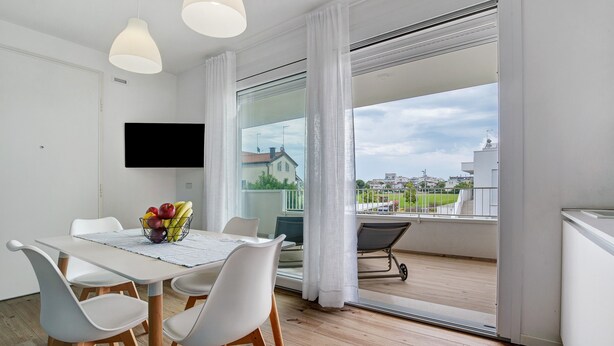 Une chambre moderne équipée d’une table à manger, de chaises, d’un téléviseur à écran plat et d’un balcon offrant une vue sur les bâtiments et la verdure.