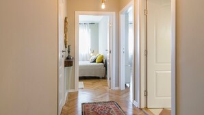 Een gang met een houten vloer die uitkomt in een slaapkamer, met een bed, nachtkastje en lamp.