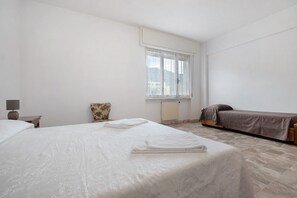 A hotel room with a large bed, a smaller bed, a chair, a desk with a lamp, and a window with a view of buildings.