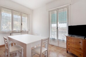 A room with a white table, four chairs, a wooden dresser, a television, and a sliding glass door with a patterned curtain.