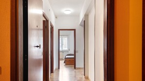 Un pasillo con una cama a lo lejos, una puerta a la izquierda y una ventana con cortinas.
