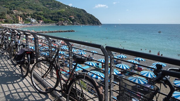 Una playa con bicicletas estacionadas a lo largo del paseo marítimo, sombrillas y vistas al mar y a las colinas.