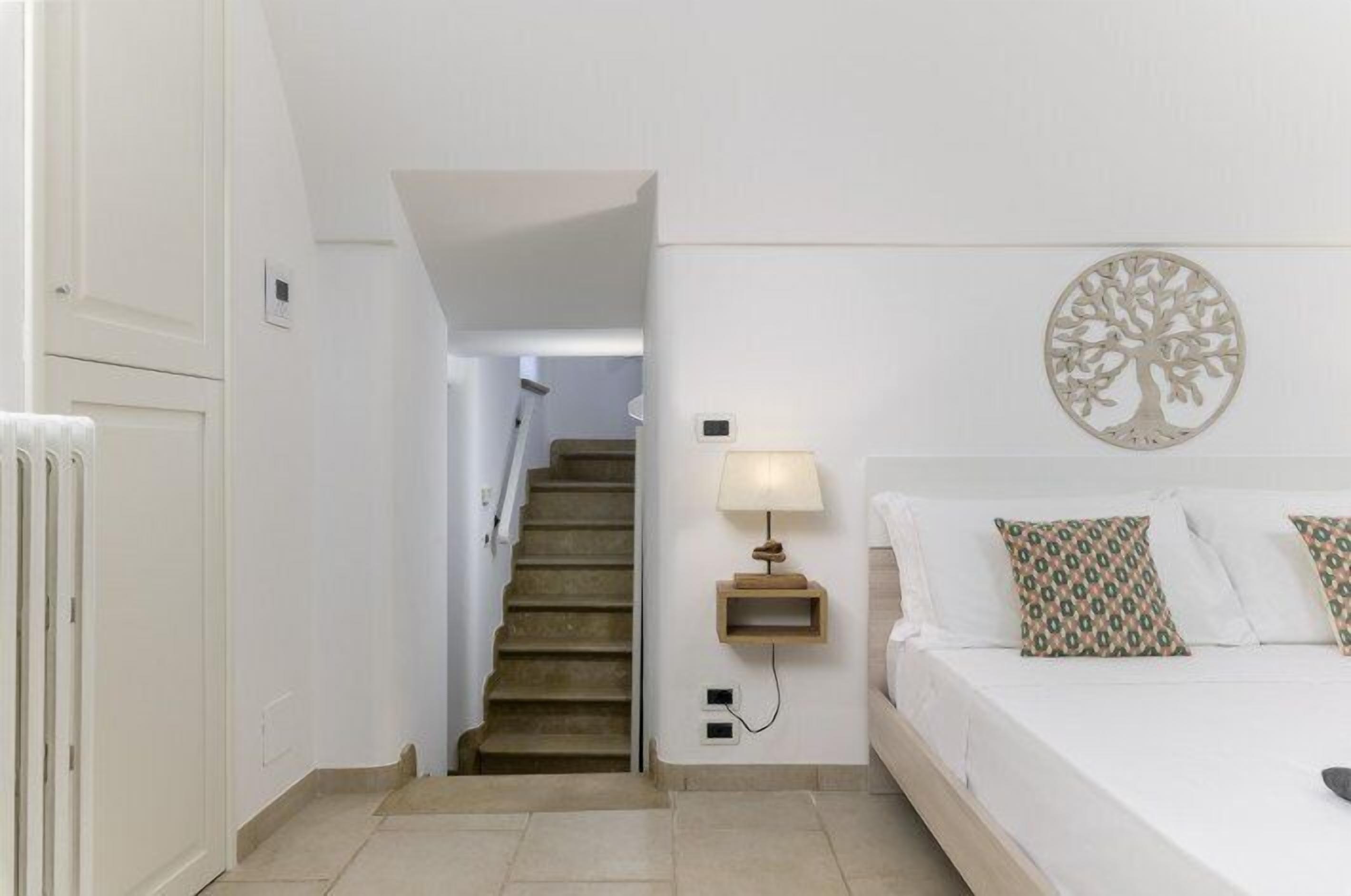 A bedroom with a bed, a nightstand, a wall-mounted lamp, a staircase, and a decorative tree wall art piece.