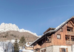 A wooden chalet with a stone foundation, a balcony, and a view of snow-covered mountains.