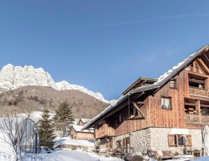 Un chalet en bois, avec des fondations en pierre, un balcon et une vue sur des montagnes enneigées.