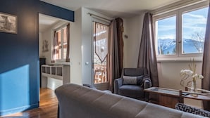 A living room with a sofa, a chair, a table with a vase, and a view of mountains through a window. - Apartment 'Les Lutins' with Mountain View, Shared Terrace and Wi-Fi (Saint-Ferréol)
