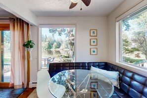 A modern living room with a glass table, black leather sofa, and large windows offering a view of a lake and trees. - Family-friendly lakefront escape with outdoor dining, gas grill, kayaks and W/D (Lacey)
