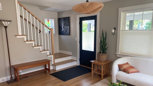 A modern living room with a staircase, a sofa, a coffee table with a plant, a hanging light fixture, and a door with a view outside. - Short Walk to the Big House, Modern Spacious 3 BR + Basement Suite, 3.5 baths (Ann Arbor)