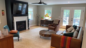 A living room with a fireplace, a television, a ceiling fan, and a view of the outdoors through large windows. - Short Walk to the Big House, Modern Spacious 3 BR + Basement Suite, 3.5 baths (Ann Arbor)
