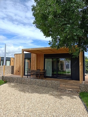 A modern house with a wooden deck, a dining area, and a large glass door leading to a covered patio. - Tidy Chalet With Dishwasher, 9 km From Maastricht (Berg en Terblijt)