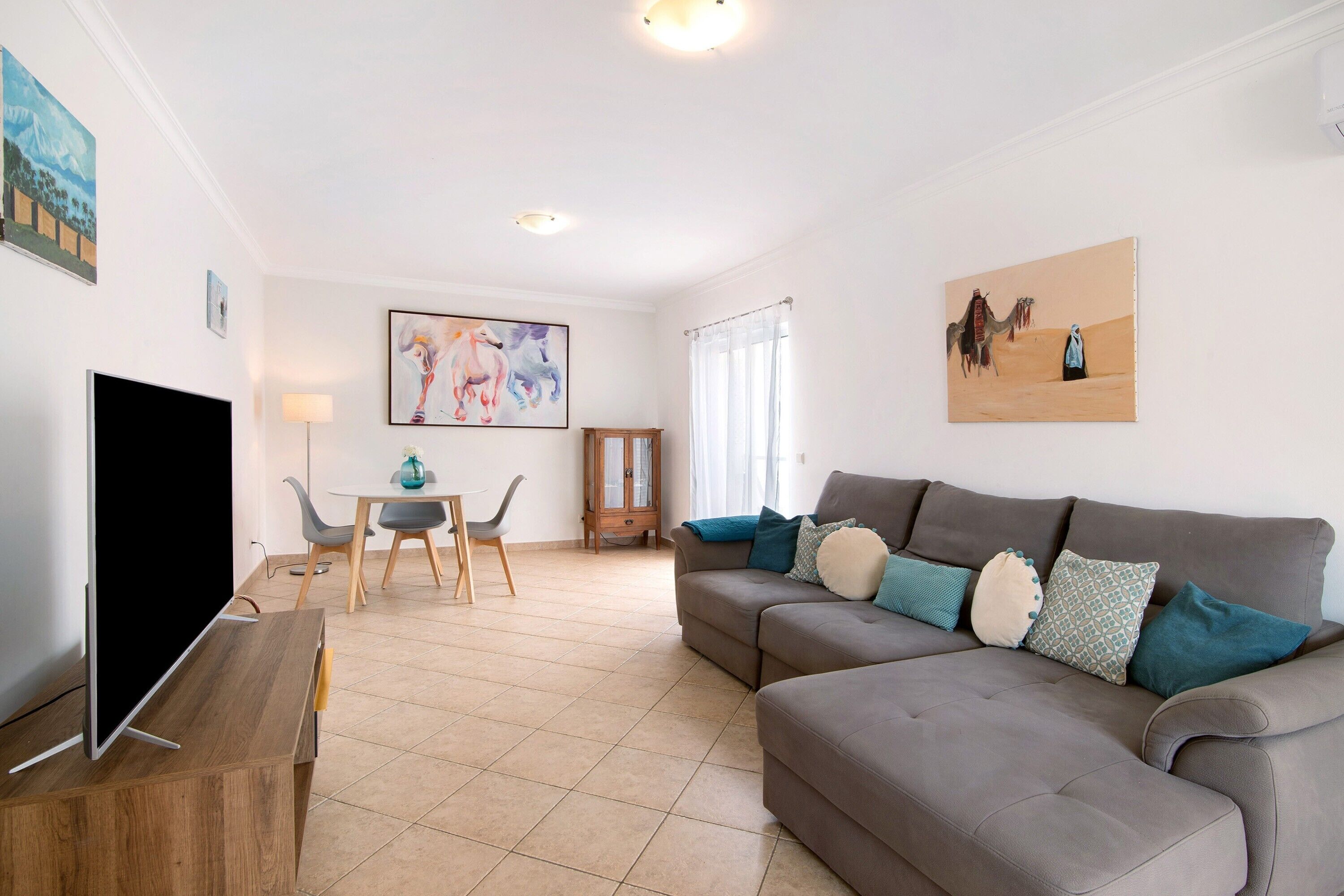 Ein Wohnzimmer mit Flachbildfernseher, grauem Sofa mit gemusterten Kissen, Holztisch und einem Bild an der Wand.