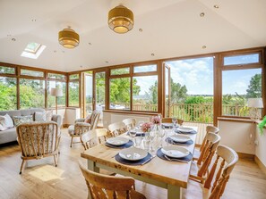 A dining area with a wooden table set for six, surrounded by wooden chairs, large windows offering a view of greenery, and a ceiling with recessed lighting. - 4 bedroom accommodation in Swansea (Swansea)