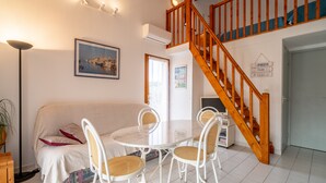 Ein Wohnzimmer mit einem weißen Sofa, einem runden Tisch und vier Korbstühlen. Eine Holztreppe mit blauer Geländer führt zu einer oberen Ebene.