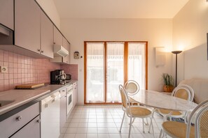 A kitchen with white cabinets, a dining table with white chairs, and a sliding glass door leading to an outdoor area. - Villa Gruissan Seaside- Les Ayguades (Gruissan)