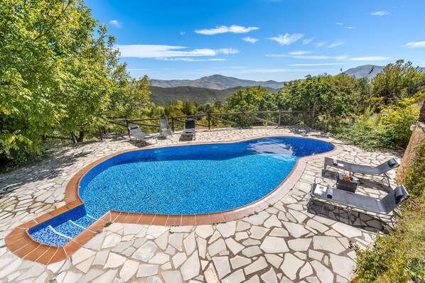 A swimming pool with a stone patio, lounge chairs, and a mountainous backdrop.