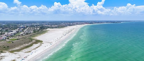 An aerial view of a coastal city with a long beach, clear blue sky, and scattered clouds.