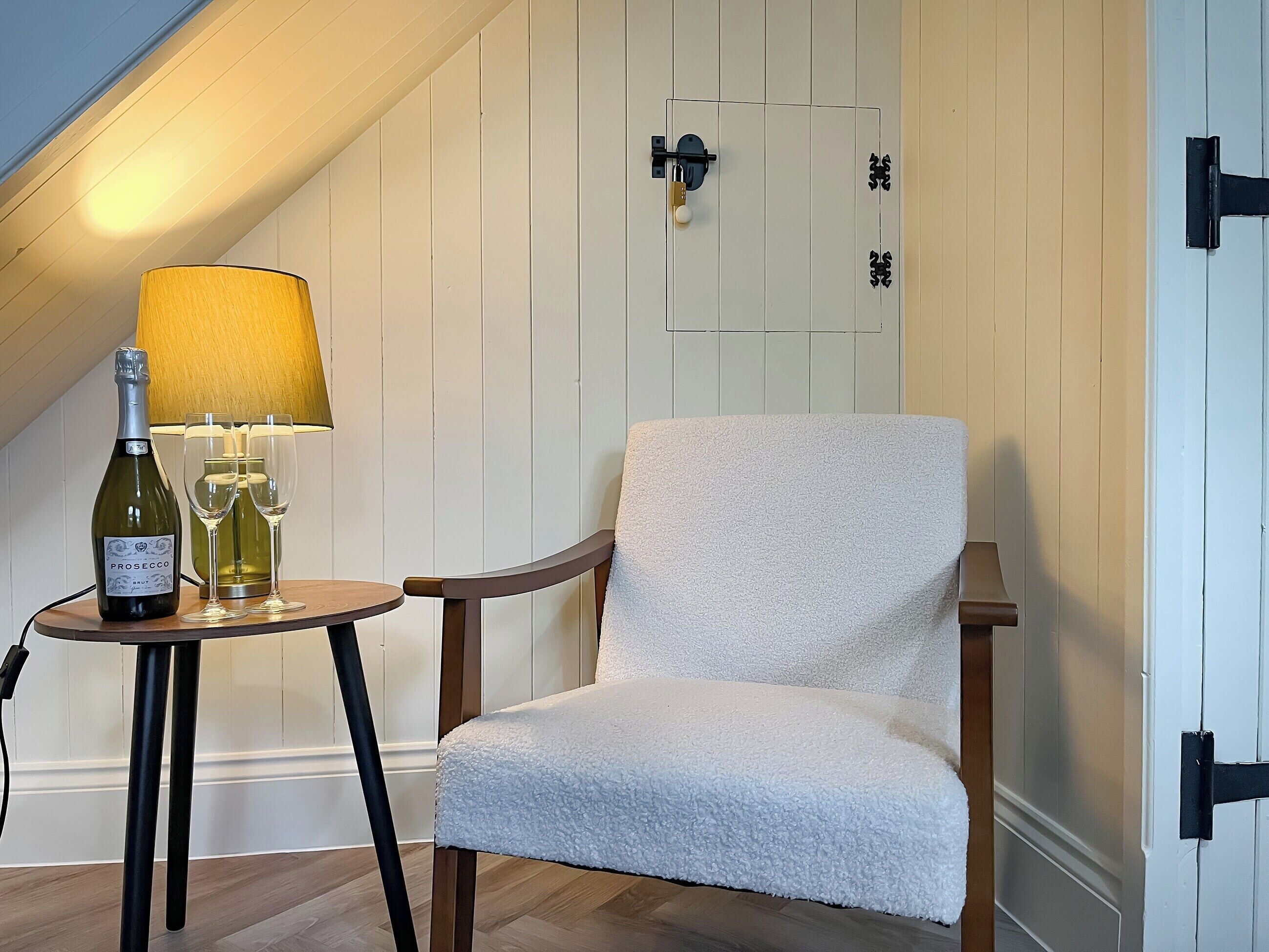 A cozy room with a white upholstered chair, a small round table with a bottle of wine and glasses, and a lamp.