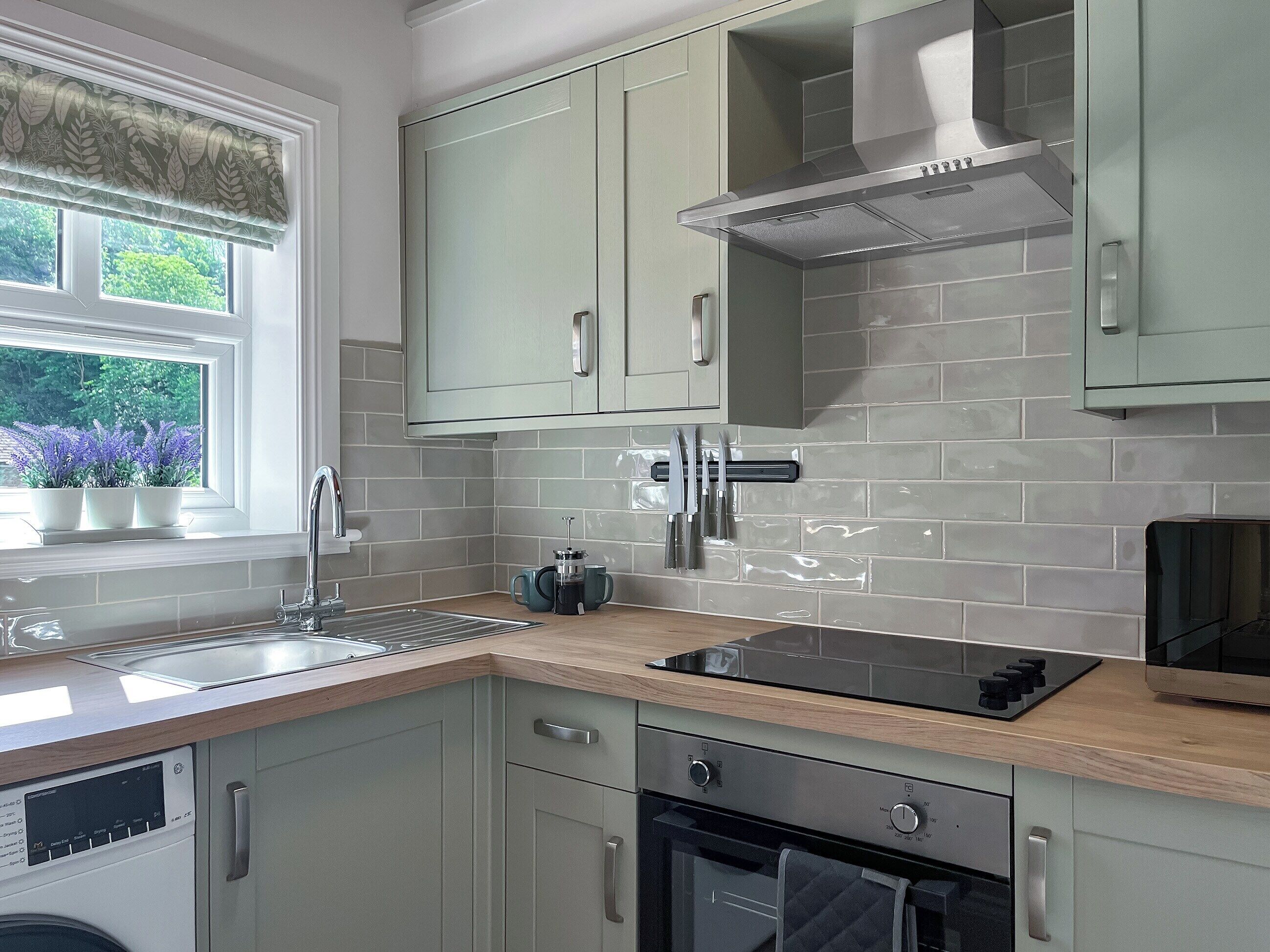 A modern kitchen with light green cabinets, a stainless steel range hood, a built-in oven, and a window with a view of greenery.