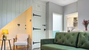 Un soggiorno con un divano verde, una poltrona bianca, un tavolino con una bottiglia e dei bicchieri, e una scala che conduce a una zona cucina.