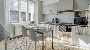 A modern kitchen with a dining table, chairs, a window with blinds, a stove, and a refrigerator. - Holiday Home 'Maison Chaleureuse Cancale' with Private Terrace and Garden (Cancale)