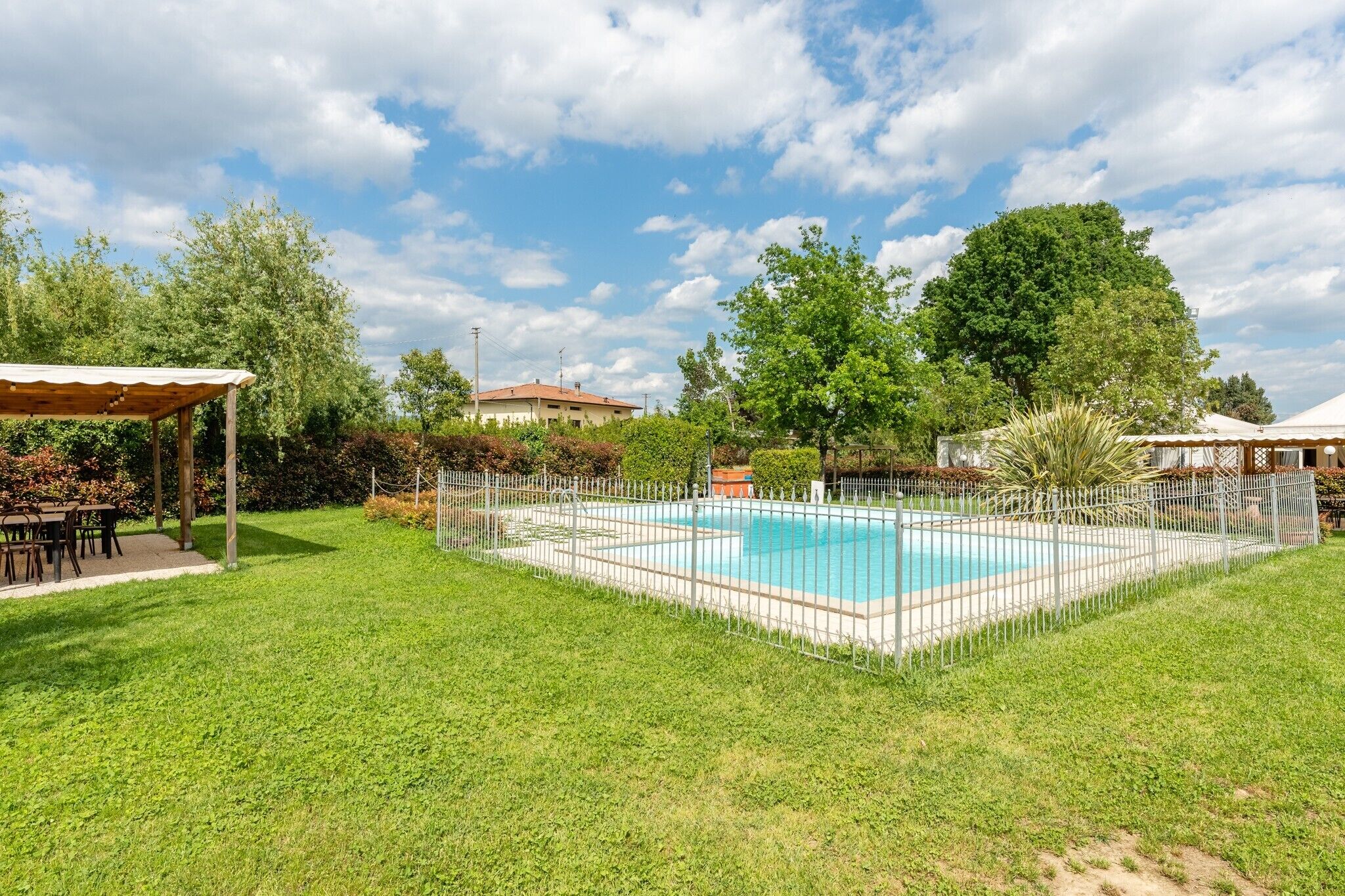 A swimming pool surrounded by a white fence, with a covered seating area and lush greenery.