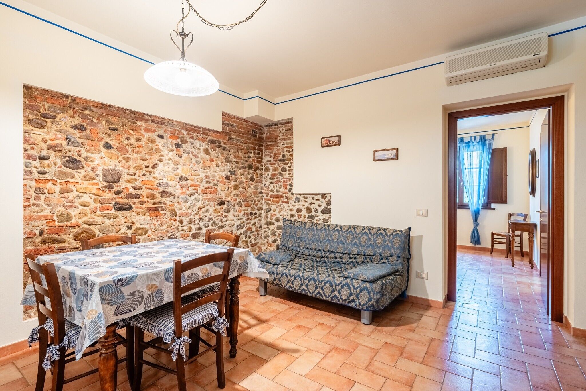 A dining area with a stone wall, a table with a patterned tablecloth, wooden chairs, and a blue patterned sofa.