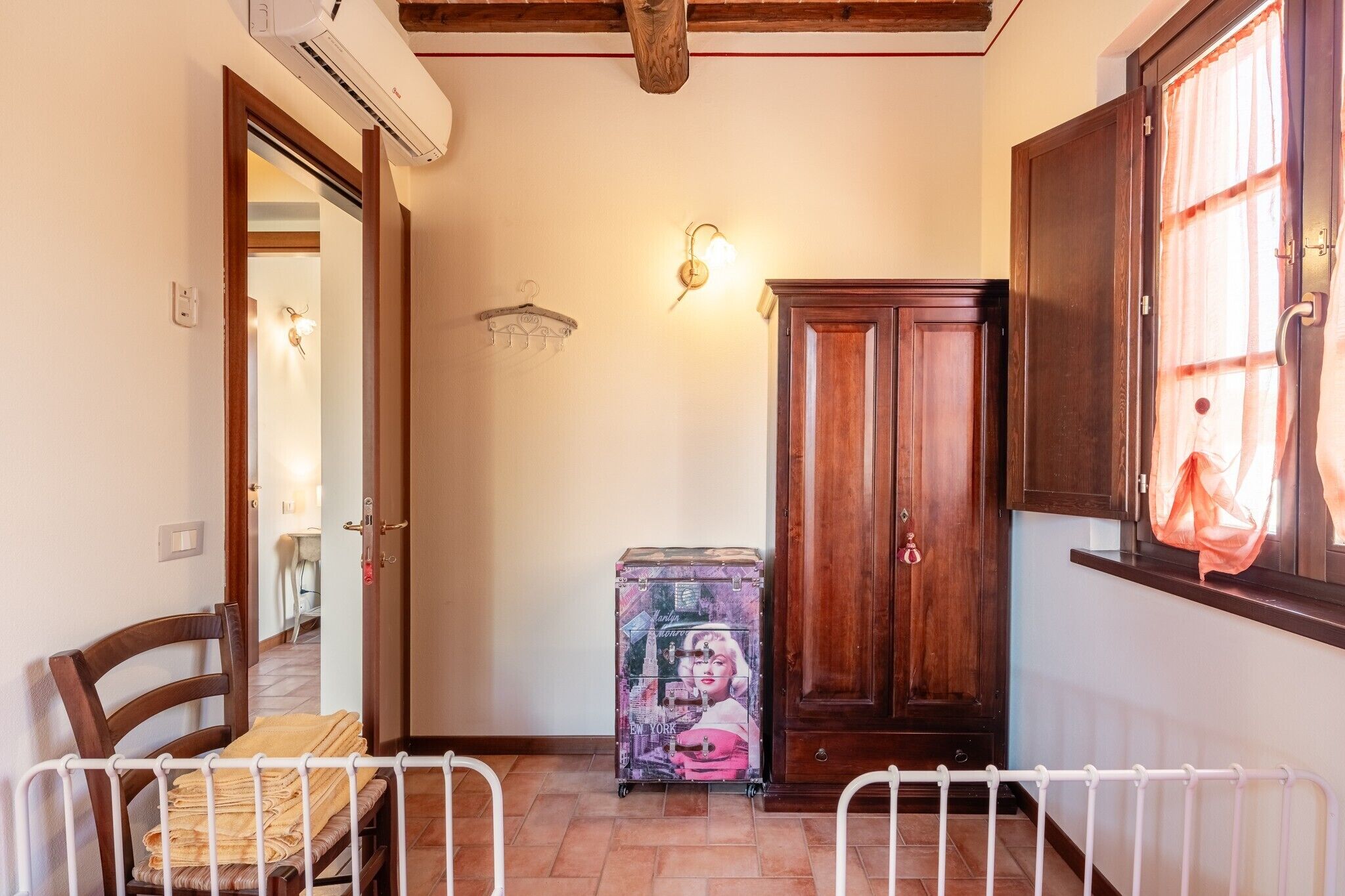 Una habitació amb un armari de fusta, una maleta, una cadira i un llit amb estructura de metall blanca.