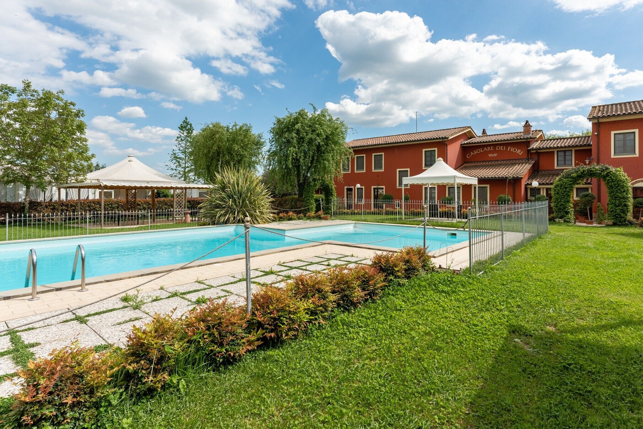 Una piscina amb una zona enjardinada tancada, un cobert blanc i un edifici vermell amb el rètol 'CASOLE DEI Fiori'.