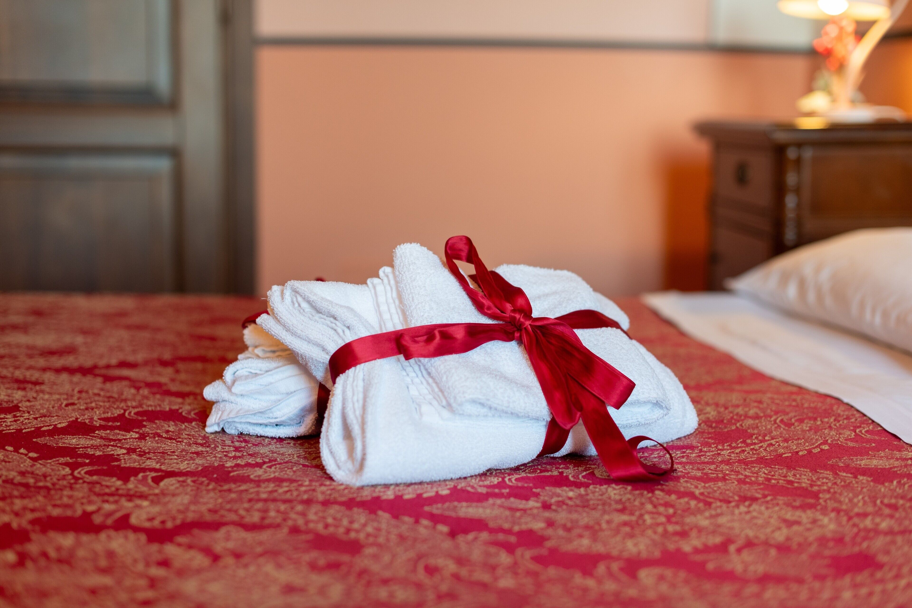 A neatly folded set of white towels tied with a red ribbon on a bed with a red patterned bedspread.